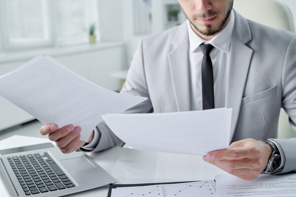 Hombre joven revisando unos documentos impresos con una MAC del lado derecho