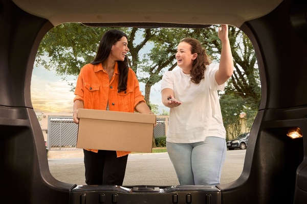 Dos mujeres sonriendo, de pie, frente a la cajuela de una de ellas